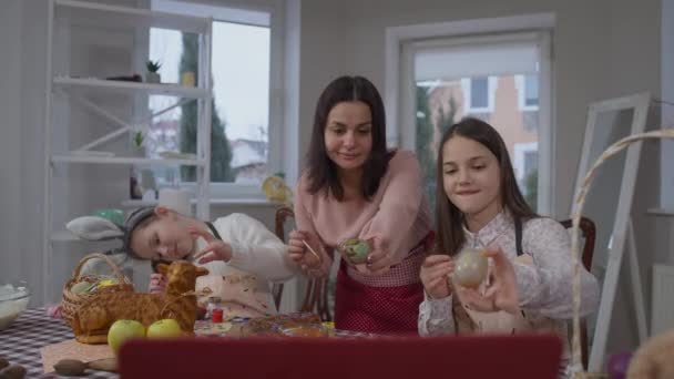 Весела мати і доньки плескають кольорові яйця на Великдень онлайн, використовуючи відеочат на ноутбуці. Позитивна кавказька жінка і дівчата показують багатокольорові страви, що посміхаються, спілкуючись у соціальних мережах.. — стокове відео