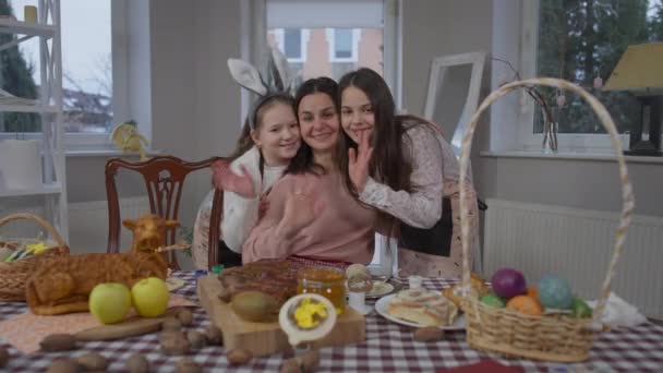Sebevědomá šťastná matka a dcery mávající kamerou, usmívající se a objímající se doma o Velikonocích. Pohled zepředu portrét veselé krásné ženy a dívek pózujících v interiéru na dovolenou. — Stock video