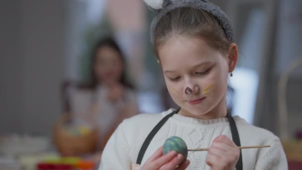 Detailní záběr okouzlující holčička pózuje uvnitř zbarvení velikonoční vejce. Roztomilé bělošské dítě hledící na kameru s namalovanou tváří. Životní styl a koncept dětství. — Stock video