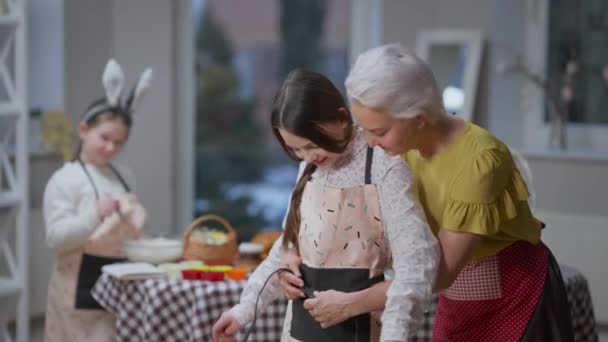 Leende vacker mormor binda förkläde på tonåring barnbarn med liten flicka matlagning i bakgrunden. Glad vit familj förbereder påsk middag hemma. — Stockvideo