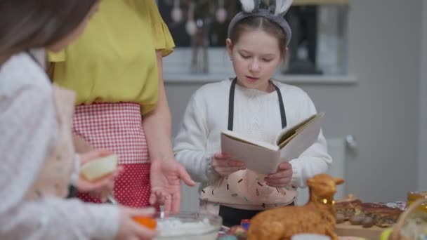 Портрет концентрированного любопытства кавказская девушка, читающая рецепт как сестра и бабушка смешивая ингредиенты на кухне дома. Красивый ребенок разговаривает помогая семье готовить пасхальный ужин в помещении. — стоковое видео
