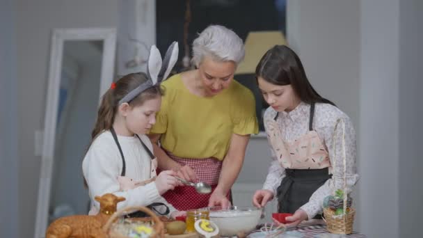 Portrait of beautiful slim grandmother talking as teenage and little granddaughters helping woman baking dessert at home indoors. Concentrated grey-haired lady and charming girls cooking on Easter. — Stock Video