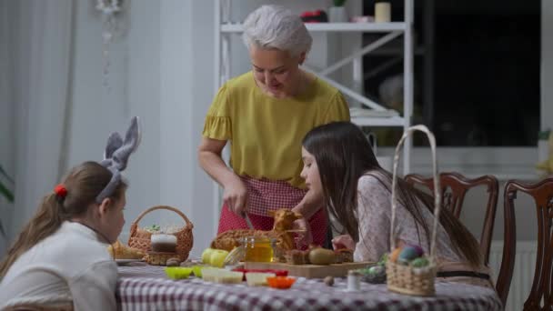 Pozytywna piękna babcia krojąc ciasto wielkanocne jagnięcina jako ciekawostki wnuczki siedzi przy stole w pomieszczeniach. Portret pewnej siebie białej kobiety tnącej smaczny słodki tradycyjny deser na wakacjach. — Wideo stockowe