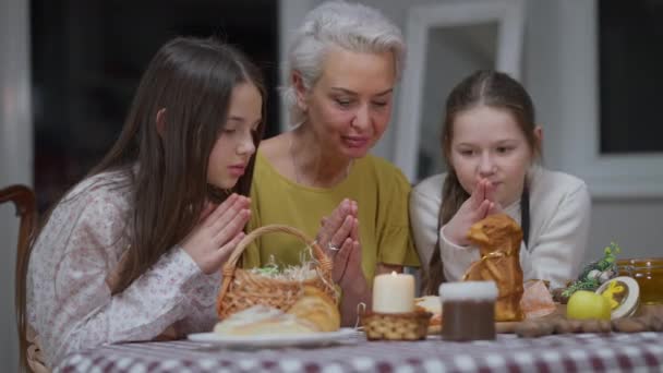 Sebevědomá běloška a dívky, modlící se o Velikonoční neděli u jídelního stolu. Pozitivní krásná babička a vnučky se sepjatýma rukama mluví děkováním Bohu. — Stock video