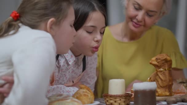 Wnuczki zdmuchują świeczkę na stole, a babcia uśmiecha się siedząc w tle. Pozytywne dziewczyny i szczęśliwa kobieta świętują Wielkanoc w domu. — Wideo stockowe