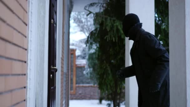 Side view cautious male burglar walking to entrance door trying to pick the lock. Young Caucasian man in black clothes looking around breaking in house on snowy winter day. Robbery and crime concept. — Stock Video