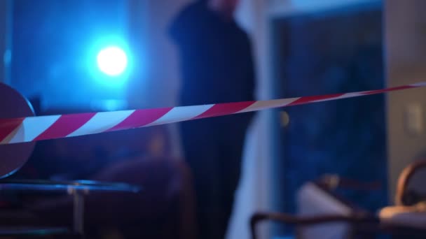 Close-up red and white crime scene barricade tape with blurred Caucasian policeman with flashlight walking at background examining upturned furniture. Concentrated cop investigating robbery. — Stock Video