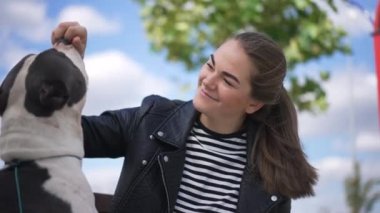 Genç ve güzel bir kadın yaz gökyüzünün arka planında beslenen bir köpekle gülümsüyor. Kafkasyalı mutlu kadın ev sahibi ve erkek evcil hayvanın hafta sonları açık havada eğlenirken portresi. Yavaş çekim.