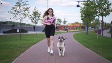 Ön manzara, şehir parkında köpekle koşan beyaz bir kadına uyuyor. Mutlu gülümseyen kadın sahibi ve erkek evcil hayvanı sabah yavaş çekimde koşuyor. Sağlıklı yaşam tarzı ve spor.