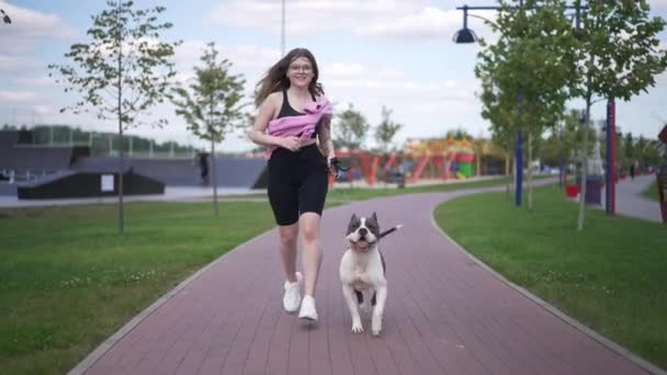 Передній вид підходить кавказькій жінці, що бігає з собакою на відкритому повітрі в міському парку. Щаслива усмішка власниці самки та тваринки бігають повільно вранці. Здоровий спосіб життя і спорт. — стокове відео
