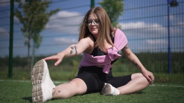 Egy motivált, pufók fiatal nő portréja, aki izmokat nyújtva melegszik fel a szabadban. Inspirált kaukázusi gyönyörű tetovált sportnő gyakorolja a napos reggel a parkban. Lassú mozgás.. — Stock videók