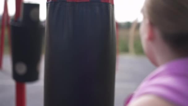 Мешок для битья, висящий на улице с татуированной женской рукой в боксёрских перчатках, пинающий оборудование в замедленной съемке. Стрельба через плечо уверенной в себе кавказской боксерской тренировки. — стоковое видео