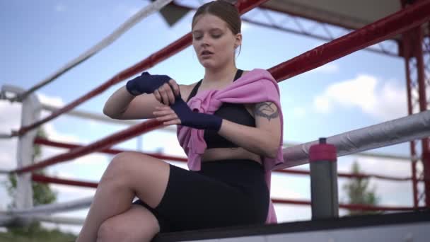 スローモーションでボクシング包帯で手首を包む美しい白人の若い女性に集中.やる気のある女性ボクサーの肖像屋外トレーニングのために準備リングに座って. — ストック動画