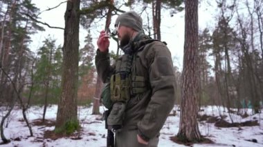 Askeri üniformalı genç beyaz adam güneş gözlüğü takıyor ve gülümseyen kameraya bakıyor. Açık havada kış ormanlarında poz veren cesur Ukraynalı bir savaşçının orta boy portresi..