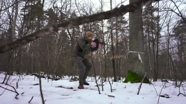 Szeroki strzał ostrożny młody człowiek w kamuflażu spacerujący z bronią wycelowaną w śnieżny zimowy las. Widok z boku Biały mężczyzna żołnierz spacerujący z bronią na zewnątrz w zwolnionym tempie. Strzał śledzący. — Wideo stockowe