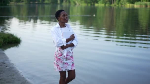 Счастливая беззаботная афро-американская молодая женщина прогуливается по берегу реки улыбаясь, глядя вверх. Портрет расслабленной довольной красивой женщины тысячелетия, идущей в замедленном движении на открытом воздухе, наслаждаясь отдыхом. — стоковое видео