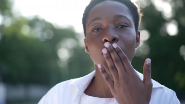 Koketa Afroameričanka gestikuluje vzduchovým polibkem a dívá se na usmívající se kameru. Detailní záběr portrét sebevědomé krásné dámy pózující ve zpomaleném filmu venku flirtování. Ženskost a krása. — Stock video