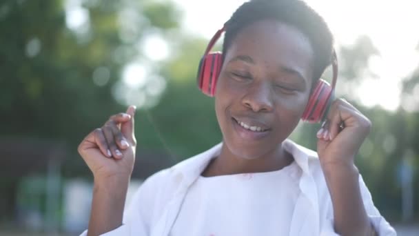 Kopfbild einer fröhlichen, unbeschwerten Afroamerikanerin, die in Zeitlupe in Sonnenstrahlen tanzt und über Kopfhörer Musik hört. Entspannt lächelnde glückliche Dame genießt Hobby am Wochenende im Park. — Stockvideo