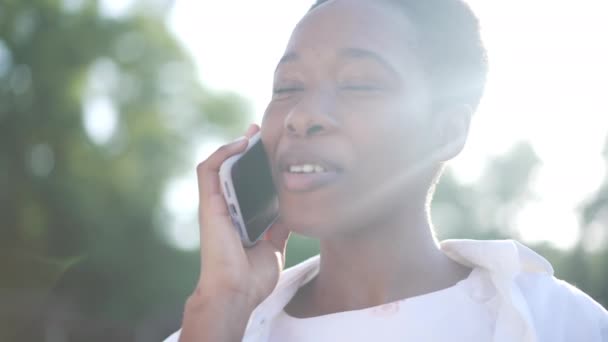 Pozitif genç Afro-Amerikan kadın telefonda konuşuyor, gülümsüyor açık havada güneş ışığında duruyor. Mutlu, kendine güvenen güzel bir bayanın yakın plan portresi.. — Stok video