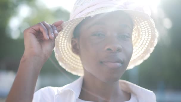 Close-up of charming African American Wanita cantik yang percaya diri dengan topi jerami Berpose di luar ruangan sinar matahari. Wanita muda tersenyum positif melihat kamera dalam gerakan lambat. Kecantikan dan feminitas. — Stok Video