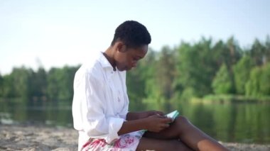 Yandan gülümseyen Afrikalı Amerikalı bir kadın kumlu nehir sahilinde oturmuş tablet üzerinde sosyal medyada sörf yapıyor. Mutlu, rahat elbiseli bayan dışarıda parkta bahar tatilinin tadını çıkarıyor. Yavaş çekim.