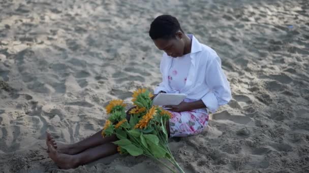 Молодая, расслабленная афроамериканская умная женщина, сидящая на песке с букетом цветов, читающая книгу. Широкий портрет умной уверенной в себе поглощенной леди, наслаждающейся хобби на песчаном пляже на открытом воздухе. — стоковое видео