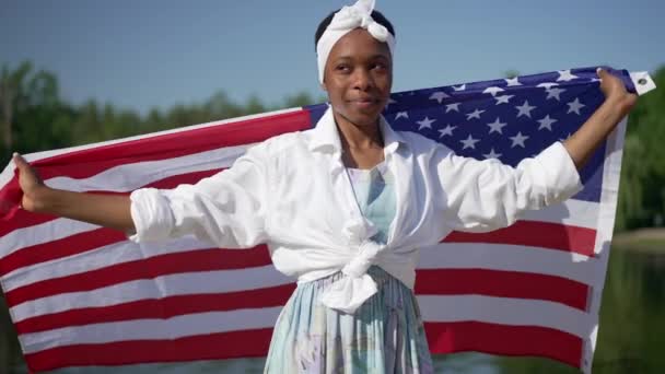 Stolze junge Afroamerikanerin, die in Zeitlupe die US-Flagge einhüllt und in die Kamera lächelt. Porträt einer glücklichen jungen schlanken Dame, die im Sommerpark hinter dem Fluss posiert. — Stockvideo