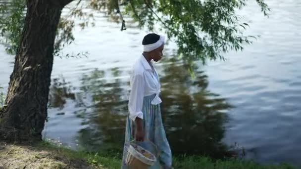 サイドビュー穏やかなアフリカ系アメリカ人の若い女性が夏の春の日に川の岸にわらバスケットで歩いています。自信を持ってスリムな女性がゆっくりと屋外で運動を散歩の撮影。自然と暮らし. — ストック動画