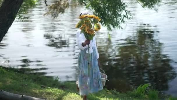 Szeroki strzał wesoła Afroamerykanka rozciągająca bukiet słoneczników uśmiechnięta patrząc na aparat podnoszący nogę w zwolnionym tempie. Portret szczęśliwej młodej damy pozującej na brzegu rzeki w zwolnionym tempie. — Wideo stockowe