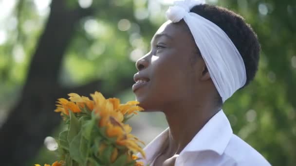 Поєднання щасливої афроамериканської жінки з букетом квітів, що милується природою, що стоїть у сонячному парку. Портрет усміхненої невимушеної жінки, яка відпочиває надворі весною.. — стокове відео