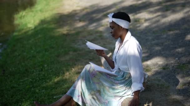 Vista lateral confiada relajada mujer afroamericana lanzando avión de papel en cámara lenta sentado al aire libre. Sonriente encantadora dama divertirse disfrutando de ocio en el parque de primavera de verano. — Vídeo de stock