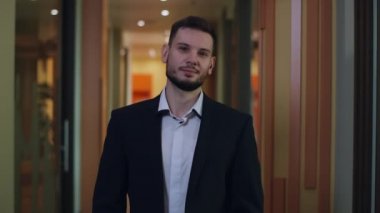 Ofis koridorunda poz veren başarılı genç beyaz iş adamı. Kahverengi gözlü ve kameraya bakıp gülümseyen esmer, yakışıklı bir adamın portresi. İş hayatı.