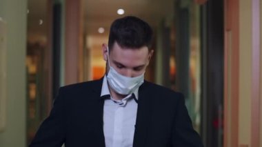 Takım elbiseli ve ofis koridorunda poz veren kendinden emin genç iş adamı. Yakışıklı beyaz adam iş merkezinde duruyor ve Coronavirus virüsü salgını sırasında kameraya bakıyor..