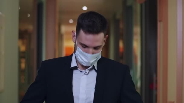 Joven empresario de confianza con traje y máscara facial Covid posando en el pasillo de la oficina. Hombre caucásico guapo de pie en el centro de negocios y mirando a la cámara durante la pandemia viral coronavirus. — Vídeo de stock