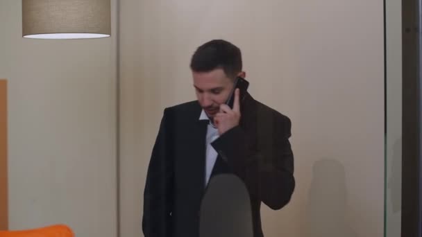Porträtt av nervös ung man som pratar i telefon på glashytten. Koncentrerad kaukasisk affärsman i kostym diskuterar projekt. Allvarlig manlig anställd på arbetsplatsen. Affärsidé. — Stockvideo