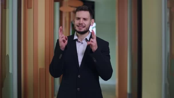 Retrato de plano mediano del hombre caucásico en traje de pie en el pasillo de la oficina con los dedos cruzados mirando hacia arriba. Guapo joven empleado masculino con la esperanza de éxito del proyecto. — Vídeos de Stock