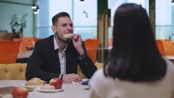 オフィスで昼食をとっている白人のビジネス関係者。ハンサムな若いビジネスマンの肖像甘いドーナツを食べ、ブルネットのビジネスマンの女性が話しているのを聞く。ライフスタイルコンセプト. — ストック動画