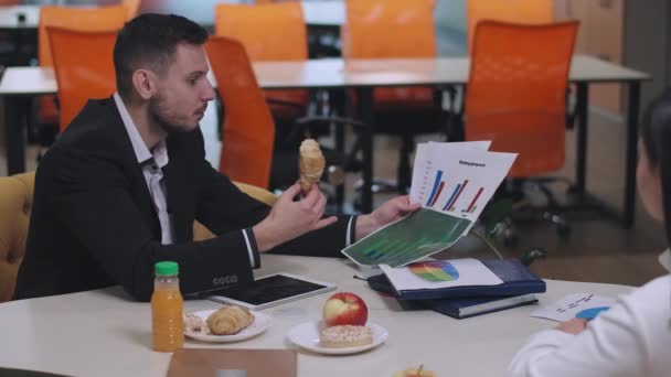 Zrelaksowany, biały biznesmen jedzący pączki i rozmawiający z kolegą podczas przerwy w pracy. Portret przepracowanego młodzieńca prezentujący diagramy współpracownikowi lub partnerowi biznesowemu podczas lunchu. — Wideo stockowe