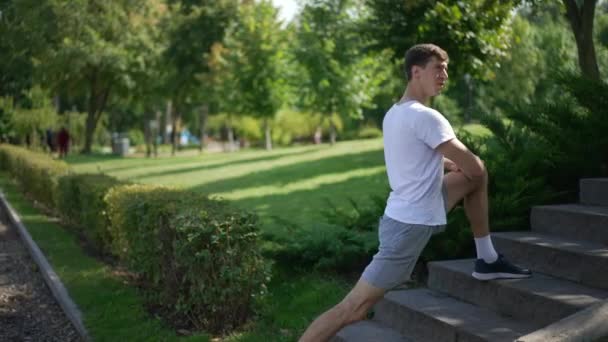 Side view young man stretching leg muscles in slow motion outdoors. Concentrated inspired Caucasian sportsman warming up in spring summer park. Workout and healthy lifestyle. — Stock Video
