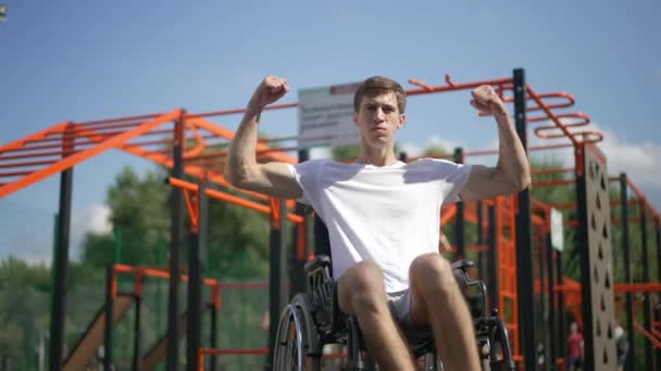 Young positive man in wheelchair gesturing strength smiling looking at camera. Portrait of motivated confident Caucasian person with disability posing on sunny sports ground outdoors in park. — Stock Video