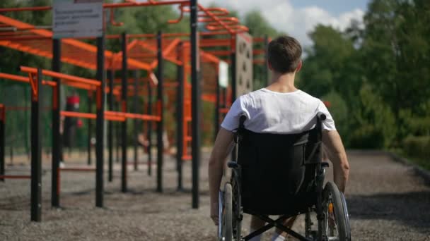 Back view young man rolling wheelchair on sports ground in spring summer park looking around. Caucasian inspired sportsman with disability riding mobility aid device in slow motion outdoors. — Stock Video