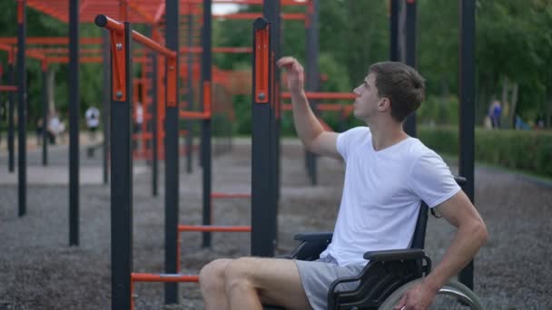 Brunetka Biały młodzieniec stojący z wózka inwalidzkiego trzymający zestaw gimnastyczny na świeżym powietrzu w parku. Motywowany sportowiec z niepełnosprawnością ćwiczy w letni wiosenny dzień. Zwolniony ruch. — Wideo stockowe