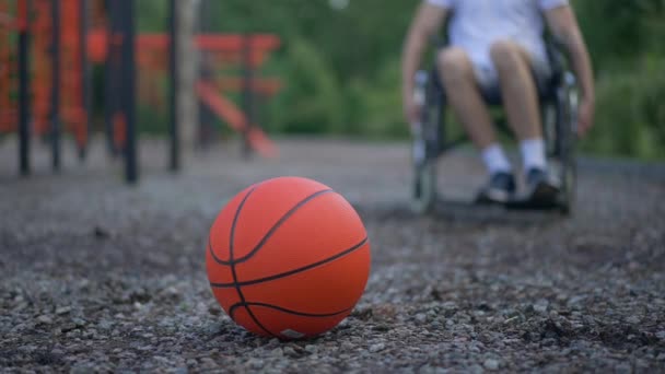 Πορτοκαλί μπάλα μπάσκετ ξαπλωμένη σε αθλητικό έδαφος, όπως ο άνθρωπος τροχαίο αναπηρική καρέκλα από εξοπλισμό ανύψωσης υποβάθρου. Νεαρός Καυκάσιος αθλητής με αναπηρία που γυμνάζεται στο πάρκο. Κίνητρο και αθλητισμός. — Αρχείο Βίντεο