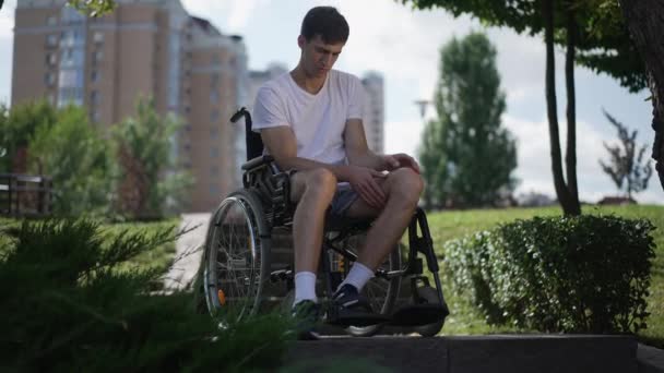Young man putting legs on wheelchair leg rest looking around thinking. Portrait of sad Caucasian guy sitting outdoors in urban city park. Disability and lifestyle concept. — ストック動画