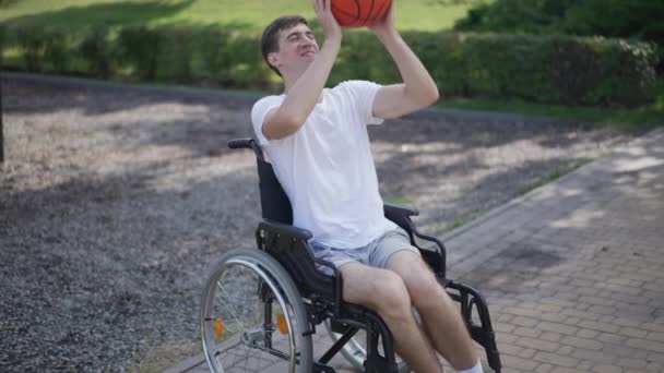 車椅子の若い陽気な白人男性の肖像屋外でスローモーションでバスケットボールボールを渡すキャッチ。晴れた日に運動を楽しむスポーツを楽しむ障害者の笑顔. — ストック動画