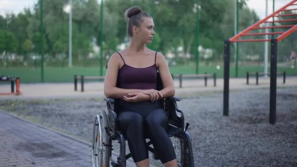 Frustrovaná krásná žena na invalidním vozíku dívá pryč sedí na sportovním hřišti v parku venku. Portrét depresivní kavkazské sportovkyně přemýšlející. Zpomalený pohyb. — Stock video