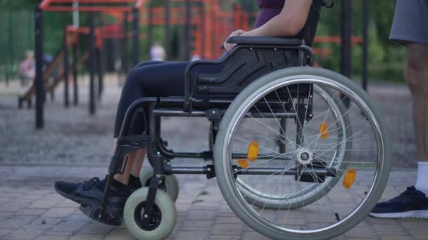 Side view unrecognizable Caucasian young woman in wheelchair with man pushing mobility aid device in slow motion leaving. Blurred sports ground in summer spring park at background. Disability concept. — ストック動画