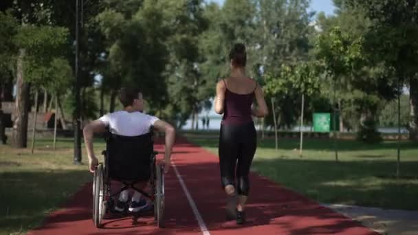 Kamera na żywo śledzi mężczyznę toczącego wózek inwalidzki jako kobieta biegająca po torze sportowym na świeżym powietrzu. Powrót widok szerokie ujęcie zmotywowanej osoby rasy kaukaskiej z niepełnosprawnością trening z dziewczyną w słoneczny dzień. — Wideo stockowe