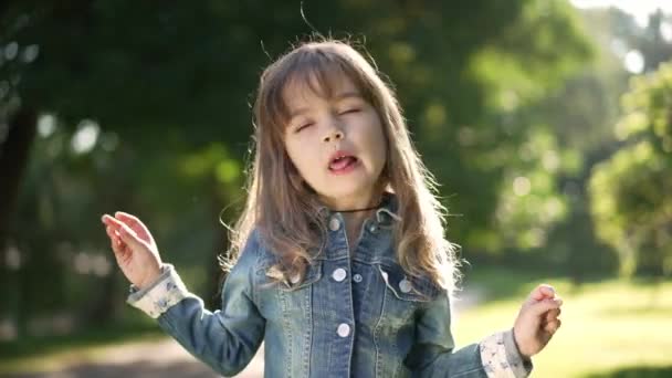 Medium shot portrait of beautiful Caucasian little girl grimacing in slow motion standing in sunny park. Pretty child making faces in sunlight outdoors. Misbehave and childhood. — ストック動画