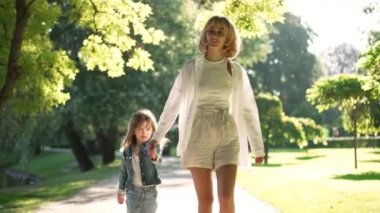 Dolly mutlu gülümseyen kadın ve kız gün ışığında el ele tutuşup konuşuyorlar. Güneşli parkta ağır çekimde gezinen muhteşem beyaz anne ve sevimli kızın ön manzara portresi.